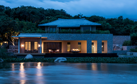 Amanoi villa with illuminated interior reflected in still water at dusk, surrounded by forested landscape.