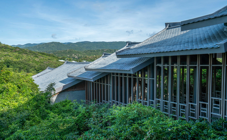 Amanoi's curved architecture extends over verdant hillside landscape beneath clear sky.