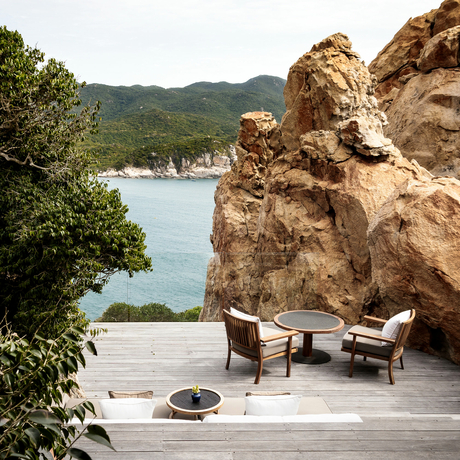 Two wooden loungers on a wooden deck beside a dramatic rocky outcrop, with calm turquoise waters and forested hills beyond at Amanoi.