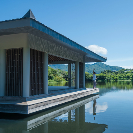 A modern pavilion with dark timber and blue-tiled roof extends over still waters at Amanoi, reflecting clear skies and distant hills.