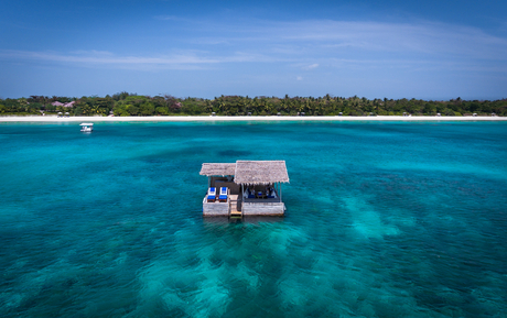 Aerial view of a wooden overwater bungalow in turquoise waters at Amanpulo, with white sand beach and tree line beyond.