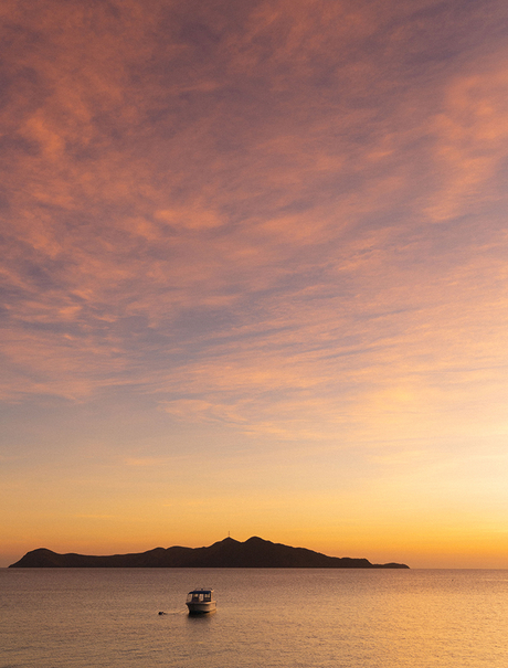 Sunset over calm waters at Amanpulo, with a small boat silhouetted against golden sky and distant island.