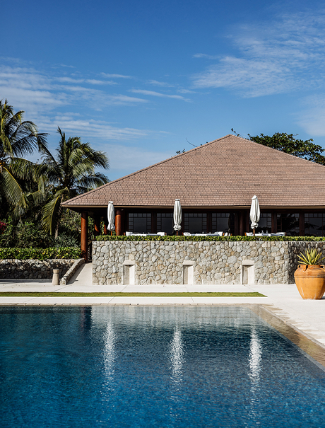 Amanpulo's open-sided pavilion with thatched roof overlooks an infinity pool beneath clear blue sky.
