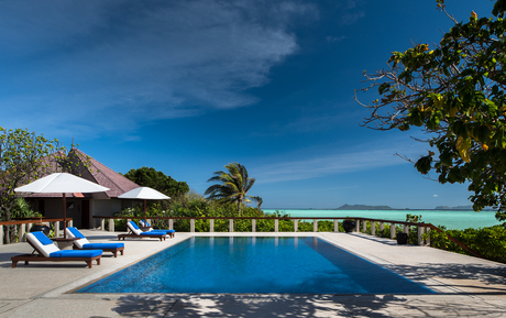 Amanpulo's swimming pool with thatched-roof pavilion and tropical gardens beneath clear blue sky.