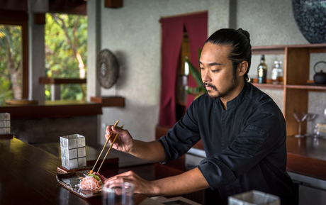 Man working at desk in Amanpulo villa interior with wooden shelving and window view.