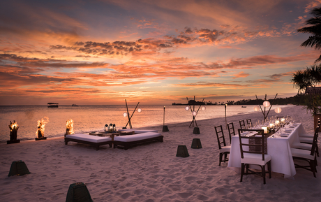 Sunset over Amanpulo's beach with moored boats and candlelit dining setup on the sand.