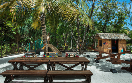 Wooden picnic table on sandy beach at Amanpulo, with palm trees and rustic building in background.