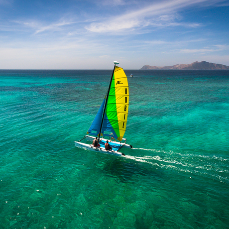 Outrigger canoe with bright yellow and green sail gliding across turquoise waters at Amanpulo.