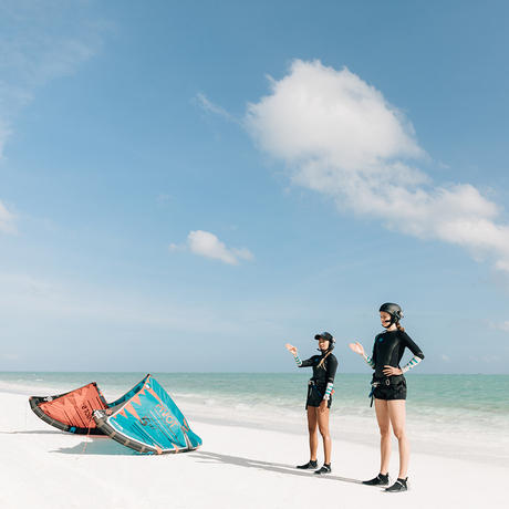 Two people stand on white sand beach at Amanpulo with kite-surfing equipment and turquoise water beyond.