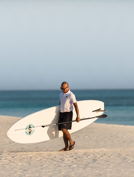 Surfer in wetsuit carrying white board on Amanpulo beach at dawn.