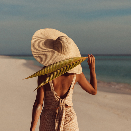 Woman wearing a wide-brimmed straw hat on a white sand beach at Amanpulo, with turquoise waters beyond.