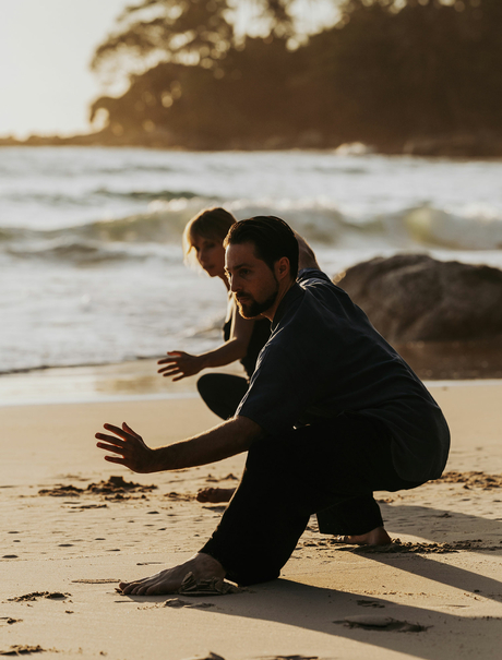 Woman in dark clothing practicing yoga on Amanpuri's beach at sunrise, waves in background.
