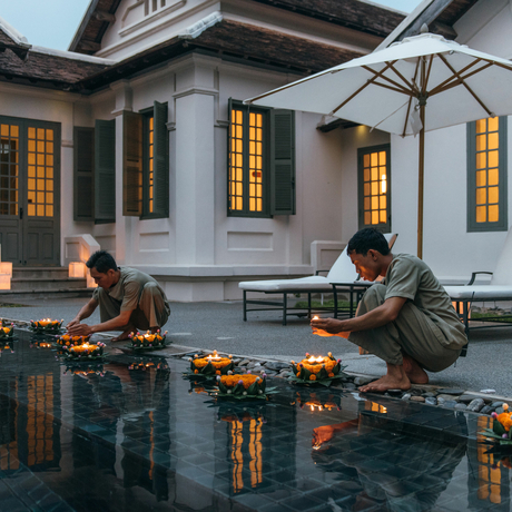 Two guests seated at opposite ends of a reflecting pool at Amantaka, with the colonial architecture and lit windows mirrored in the still water.