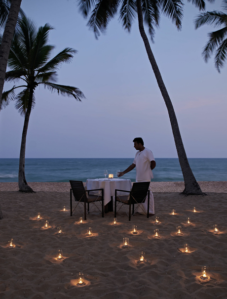 A guest sits at a dining table on Amanwella's beach at dusk, surrounded by candlelit lanterns and swaying palm trees.