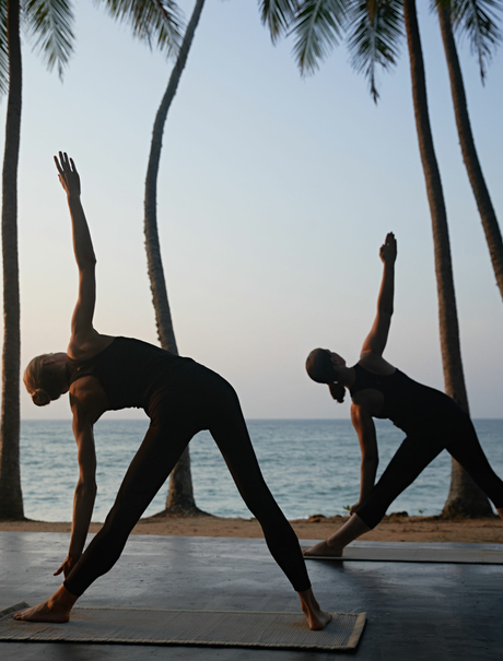 Two figures practising yoga on a wooden deck beneath palm trees overlooking the ocean at Amanwella.