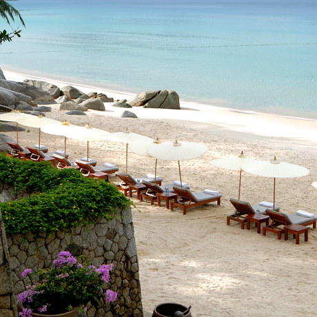 Sandy beach at Amanpuri with sun loungers beneath cream umbrellas and tropical flowers in foreground.