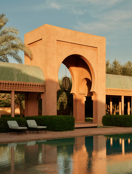 Amanjena's terracotta architecture reflected in still pool water, with palm tree and loungers visible.