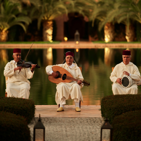 Three musicians perform at dusk on an illuminated poolside terrace at Amanjena, with palm trees reflected in the water behind them.