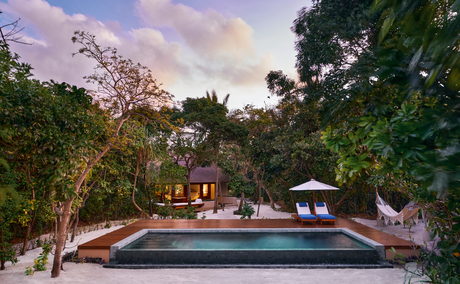 Amanpulo beach pool casita at dusk, with illuminated pavilion reflected in still water, surrounded by tropical vegetation.