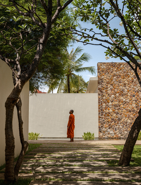 Monk in saffron robes walking through shaded courtyard at Amansara, Cambodia, framed by trees and stone walls.