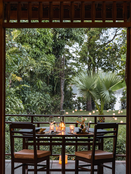 Dining table set for two on a shaded terrace at Amansara, overlooking tropical gardens and palm trees.