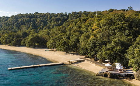 Amanwana's sandy beach meets turquoise waters, lined with dense tropical forest and traditional wooden structures.