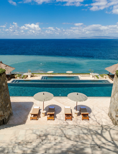 Amankila resort pool overlooking Bali's coast with white loungers and umbrellas beneath clear sky.