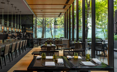 Dining venue at Aman Tokyo with floor-to-ceiling windows overlooking spring foliage, wooden ceiling beams, and dark furnishings.