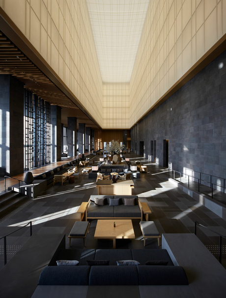 Aman Tokyo's lobby features a minimalist interior with warm wood ceiling panels, dark walls, and simple furnishings arranged along a serene corridor.