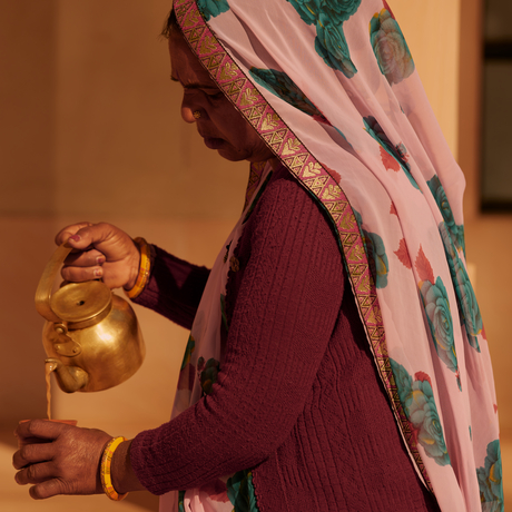 A woman at Amanbagh holds a brass watering can, wearing traditional Indian attire in deep red with a patterned shawl.