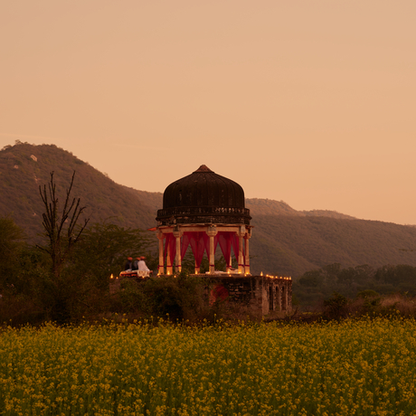 Chhatri dinner pavilion at Amanbagh, illuminated at dusk in a mustard field with mountains beyond.