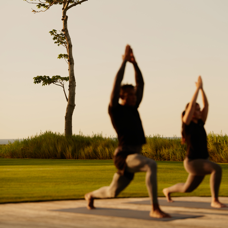 Two people practising yoga outdoors at Amanera, with manicured lawns and a single tree in the background.