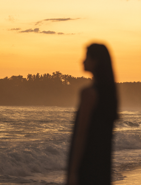 Silhouetted figure standing on Amanera's beach at sunset, golden sky reflected in calm water.