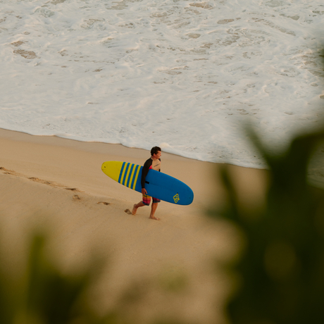 Surfer in blue wetsuit carrying yellow board walking along sandy beach at Amanera.