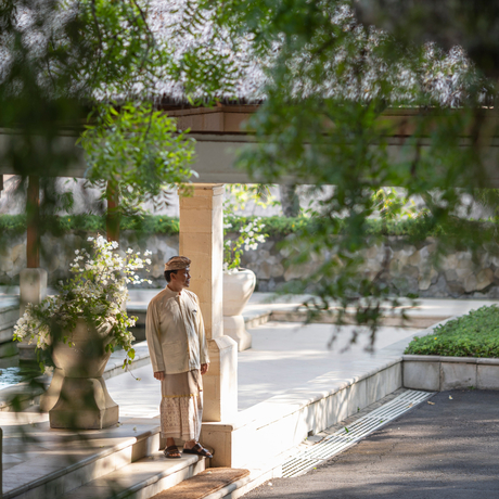 Staff member at Amankila, Indonesia, standing beneath leafy trees on a sunlit terrace.