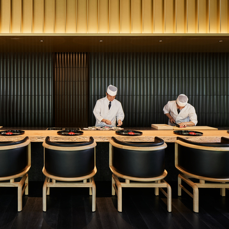Two chefs stand behind a wooden counter at Aman Kyoto, with empty dining chairs lined in front.