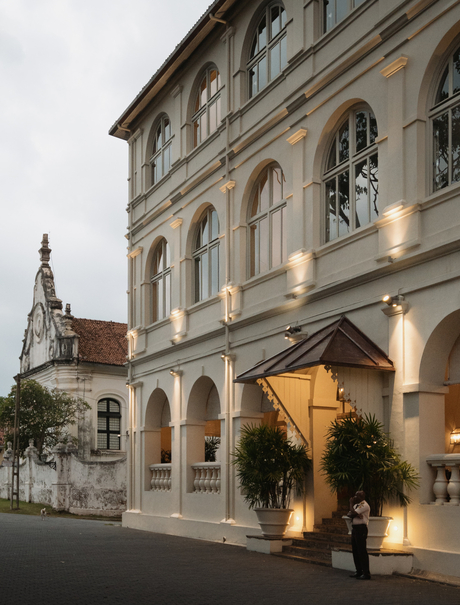 Amangalla's elegant façade at dusk, showcasing colonial architecture with illuminated ground-floor arches and a church spire beyond.