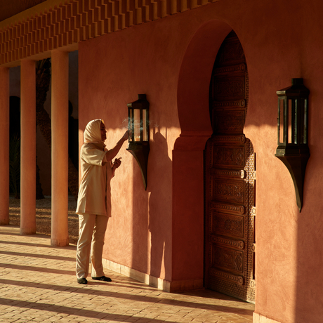 A guest stands alone on a terracotta courtyard at Amanjena, admiring the geometric shadows cast by architectural columns on the ochre-coloured wall.
