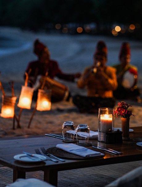 Candlelit dinner table at Amanwana with guests gathered on the beach at dusk in Sumbawa, Indonesia.