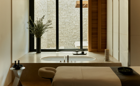 Spa treatment room at Amanzoe resort with soaking tub overlooking garden views through large windows.