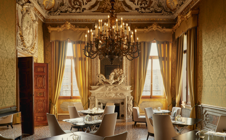 Gilded reception room at Aman Venice with ornate chandelier, classical mirrors, and tall windows draped in cream fabric.