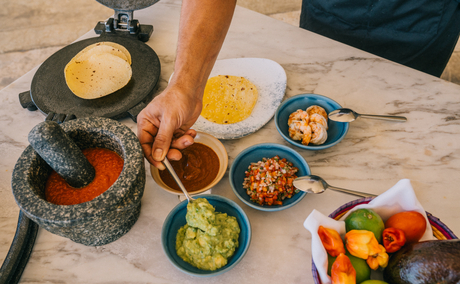 Cooking demonstration at Amanera with chef preparing dishes in multiple pans and bowls of colourful ingredients on wooden surface.