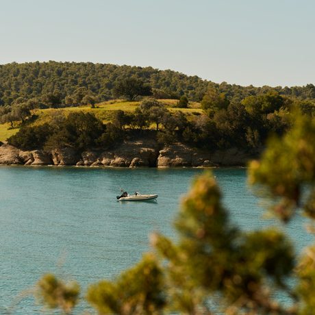 Turquoise waters and pine-covered hillside at Amanzoe beach club resort, with a small boat anchored offshore.
