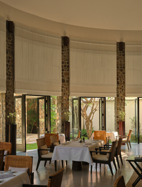 Dining room at Amansara with tall wooden columns, set table, and garden views through large windows.