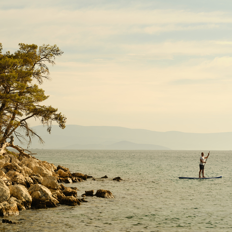 Person paddleboarding in calm turquoise waters at Amanruya, Turkey, with rocky shoreline and tree in foreground.