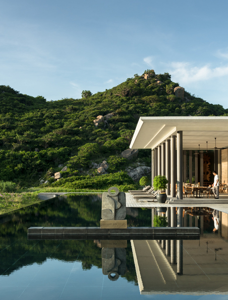 Beach club exterior at Amanoi, Vietnam, with a modern pavilion reflected in still water and forested hillside beyond.