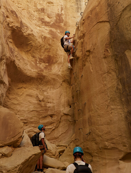 A climber scales a rust-coloured rock face during via ferrata at Amangiri, whilst two others prepare below.