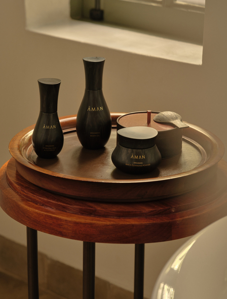Aman Essentials skincare products displayed on a wooden round table at Amantaka, featuring black bottles and a cream jar.