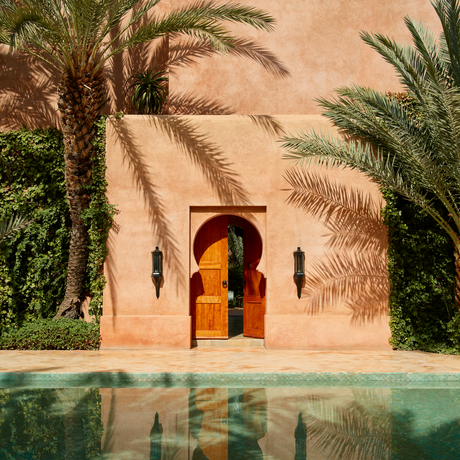 Terracotta archway with golden doors framed by palm trees and reflecting pool at Amanjena, Morocco.