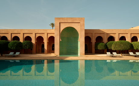 Amanjena's main pool with turquoise water reflecting the clay-coloured arched buildings and green gateway under clear sky.